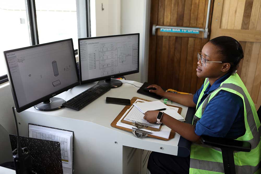 A person wearing a high-visibility vest sits at a desk with two computer monitors displaying technical diagrams. They are using a mouse and have a clipboard with papers and a pen in front of them. A wooden door with a "push bar to open" sign is in the background.